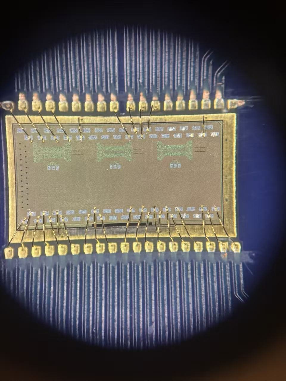 Macro close-up of a wire-bonded microchip viewed under a microscope, showing the central die with etched logic blocks and gold wire bonds running to the surrounding package contacts.